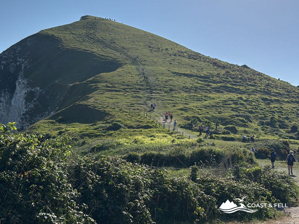 Participants climbing steep green headland during Jurassic Coast Challenge with chalk cliffs visible