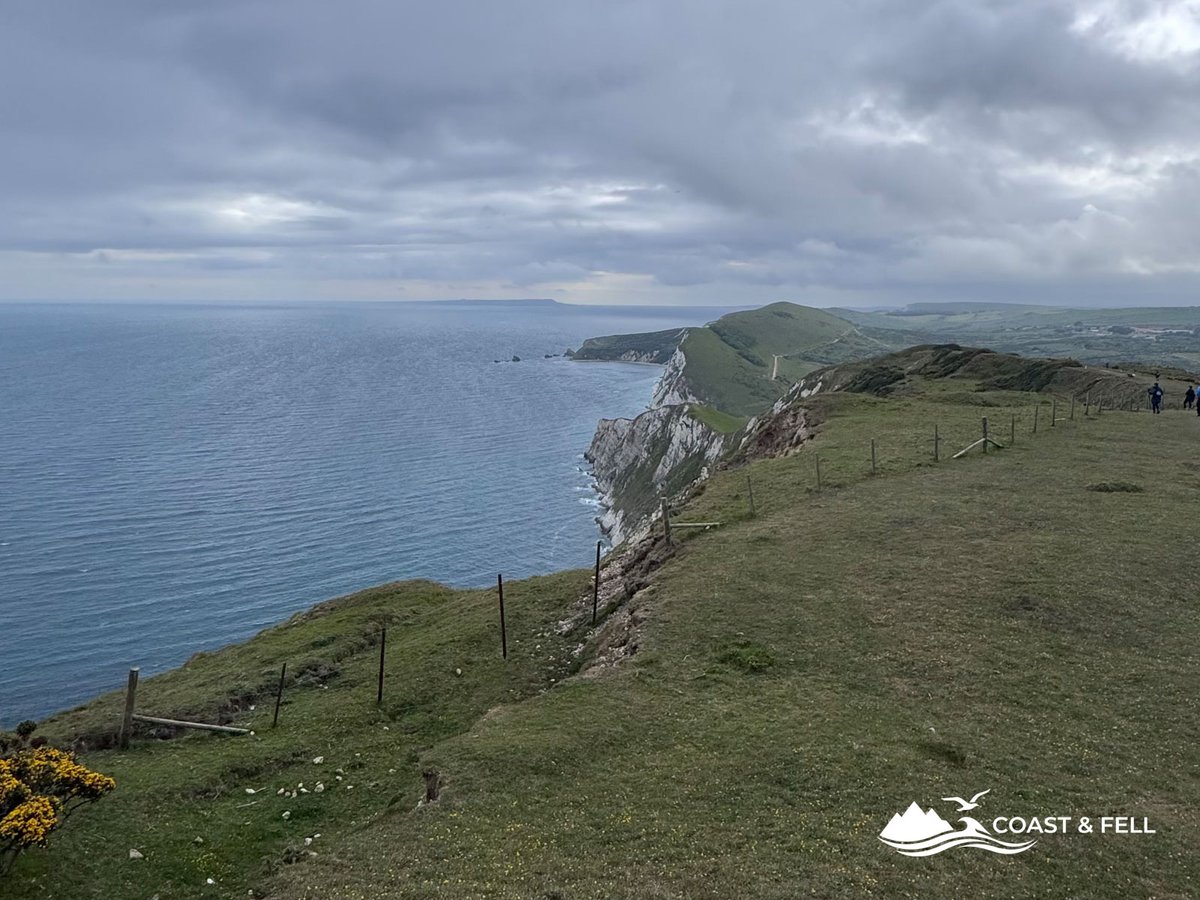 Clifftop path looking west along Jurassic Coast chalk headlands meeting grey sea under overcast sky with walkers visible in far distance
