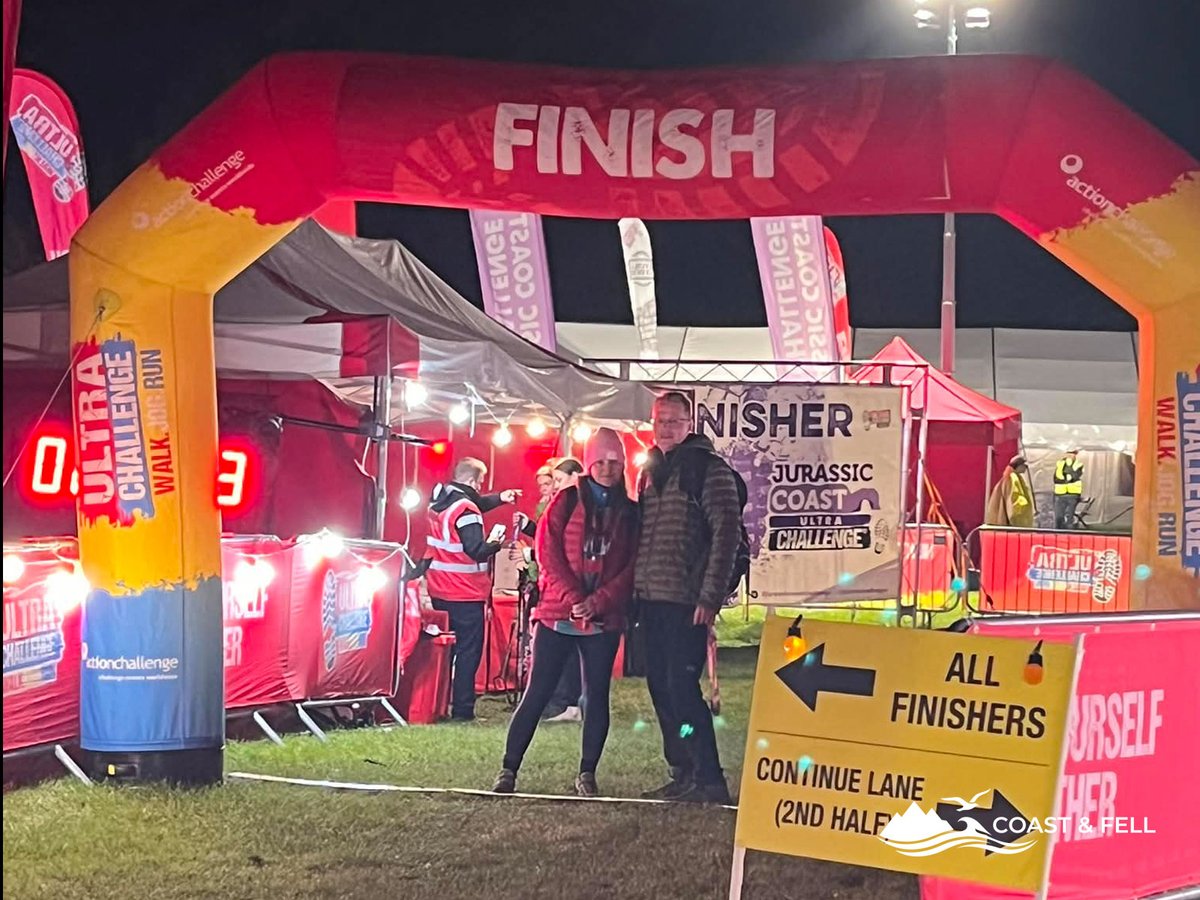 Finish arch at night for Jurassic Coast Ultra Challenge 2026 with two finishers posing in front and timing display visible